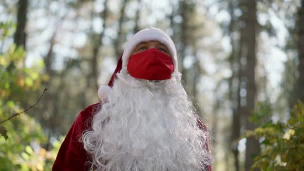 Um cara vestido com um terno de Papai Noel com uma máscara médica de proteção vermelha em seu rosto caminha sozinho na floresta. Coronavírus e Natal. Ano Novo durante a quarentena. Movimento lento — Vídeo de Stock