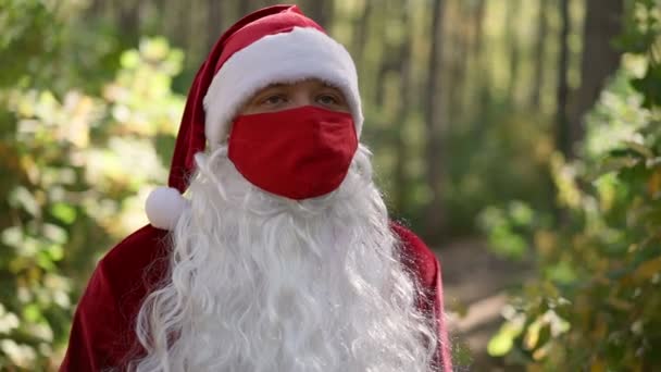 Um cara vestido com um terno de Papai Noel com uma máscara médica de proteção vermelha em seu rosto caminha sozinho na floresta. Coronavírus e Natal. Ano Novo durante a quarentena. Movimento lento — Vídeo de Stock