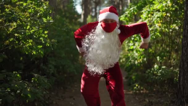 Портрет смешного человека в костюме Санта-Клауса с двумя медицинскими масками на лице стоит через лес. Рождество и коронавирус. Ковид-19. Карантин во время праздников. Медленное движение — стоковое видео