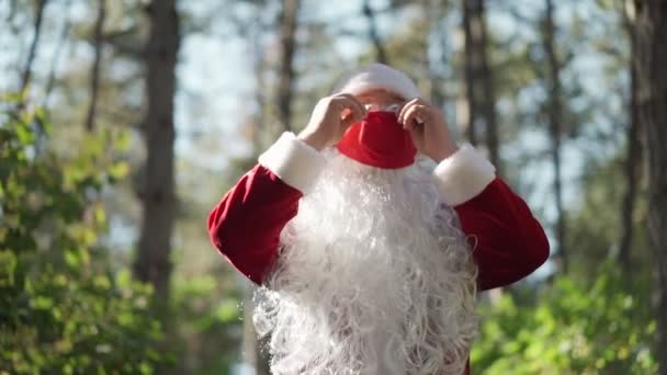 Um tipo vestido com um fato de Pai Natal põe uma máscara médica protectora vermelha na cara na floresta. Coronavírus e Natal. Ano Novo durante a quarentena. Movimento lento — Vídeo de Stock