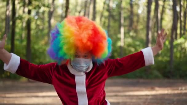 Lekfull ung kille med en clownperuk på huvudet och en medicinsk mask i ansiktet rolig dans i skogen. Coronavirus och pandemi. Karantän. Humorkoncept. Långsamma rörelser — Stockvideo