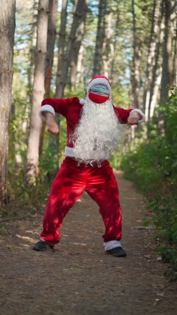 Вертикальне відео. Грайливий щасливий чоловік, одягнений у костюм Миколая з двома медичними масками на обличчі, забавно танцює в лісі. Різдво і Коронавірус Ковід-19 Карантин під час свят. Повільний рух — стокове відео