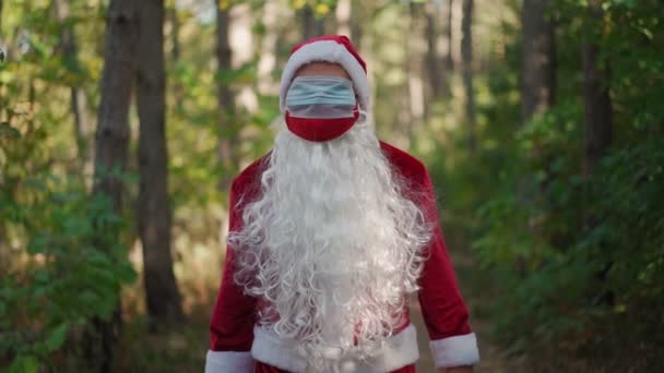 Um homem engraçado vestido com um traje de Papai Noel com duas máscaras médicas em seu rosto caminha pela floresta. Natal e coronavírus. Covid-19. Quarentena durante as férias. Movimento lento — Vídeo de Stock
