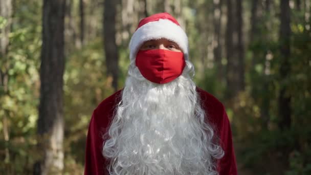 Парень в костюме Санта Клауса с защитной медицинской маской на лице стоит один в лесу. Коронавирус и Рождество. Новый год во время карантина. Медленное движение — стоковое видео