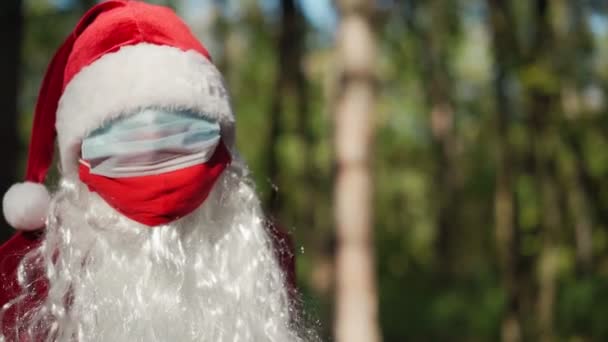 Portret zabawnego mężczyzny ubranego w kostium Świętego Mikołaja z dwoma maskami medycznymi na twarzy stoi w lesie. Święta i koronawirus. Covid-19. Kwarantanna podczas świąt. Zwolniony ruch — Wideo stockowe