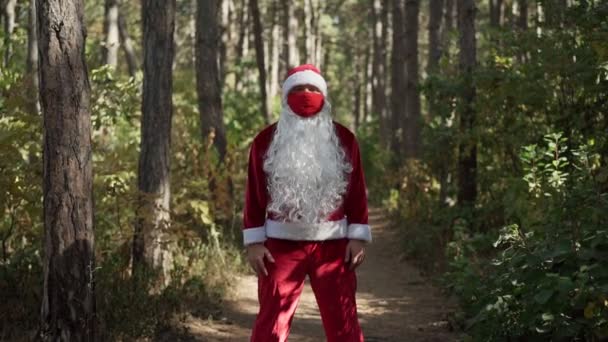 Грустный парень в костюме Санта Клауса с защитной медицинской маской на лице стоит один в лесу. Коронавирус и Рождество. Новый год во время карантина. Медленное движение — стоковое видео