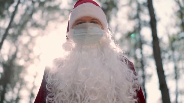 Un gars vêtu d'un costume de Père Noël avec un masque médical de protection sur le visage se tient seul dans la forêt par une journée ensoleillée. Coronavirus et Noël. Nouvel An en quarantaine. Mouvement lent — Video