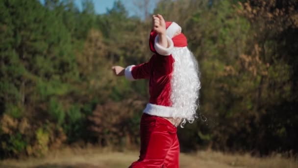 Brincalhão homem feliz vestido em terno Papai Noel com máscara médica em seu rosto engraçado dançando na floresta. Natal e coronavírus Covid-19 Quarentena durante as férias. Movimento lento — Vídeo de Stock