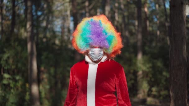 En ung rolig kille med en clownperuk på huvudet och en medicinsk mask i ansiktet i skogen. Coronavirus och pandemi. Karantän. Humorkoncept. Långsamma rörelser — Stockvideo