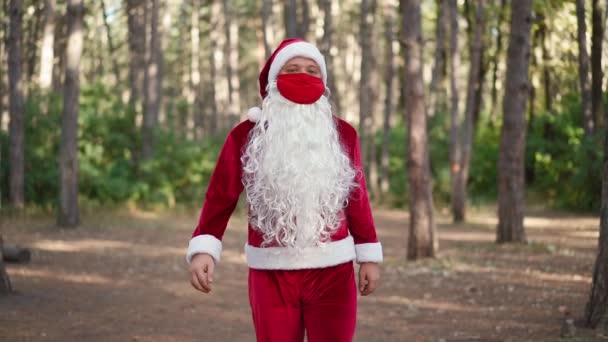Um cara vestido com um terno de Papai Noel com uma máscara médica de proteção vermelha em seu rosto caminha sozinho na floresta. Coronavírus e Natal. Ano Novo durante a quarentena. Movimento lento — Vídeo de Stock