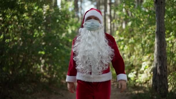 Um homem engraçado vestido com um traje de Papai Noel com máscara médica em seu rosto caminha pela floresta. Natal e coronavírus. Covid-19. Quarentena durante as férias. Movimento lento — Vídeo de Stock