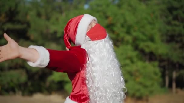 Игривый счастливый человек, одетый в костюм Санта-Клауса в медицинскую маску, забавно танцует в лесу. Рождественский и коронавирус Ковид-19 Карантин в праздничные дни. Медленное движение — стоковое видео