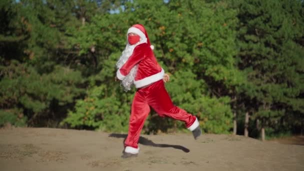 Játékos boldog ember öltözött Mikulás öltöny védő orvosi maszk az arcán vicces tánc az erdőben. Karácsony és koronavírus Covid-19 Karantén az ünnepek alatt. Lassú mozgás. — Stock videók
