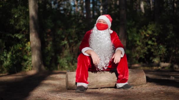 Verdrietige eenzame man gekleed in een kerstman pak met een beschermend medisch masker op zijn gezicht zittend op een boomstronk alleen in het bos. Coronavirus en Kerstmis. Nieuwjaar tijdens quarantaine. Langzame beweging — Stockvideo