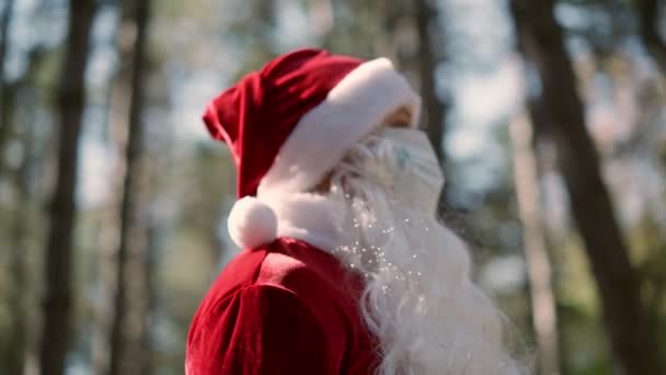 En kille klädd i tomtedräkt med en skyddande medicinsk mask i ansiktet står ensam i skogen en solig dag. Coronavirus och jul. Nytt år under karantänen. Långsamma rörelser — Stockvideo