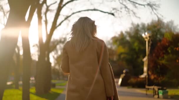 Oigenkännlig vacker affärskvinna i en brun rock promenader genom stadsparken för att arbeta på kontoret på hösten i solnedgången ljus. Bakåt. Långsamma rörelser — Stockvideo
