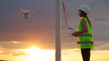 Üniformalı ve kasklı kadın ekoloji mühendisi elinde joystick ile yel değirmeninde çalışan uçan dronu kontrol ediyor. Elektrik enerjisine alternatif. Yavaş çekim.