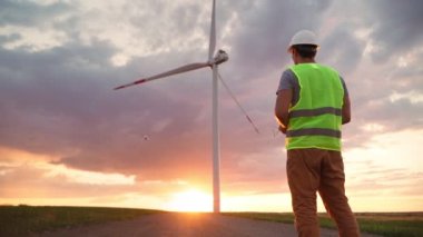 Üniformalı ve miğferli Adam Profesyonel Ekoloji Mühendisi elinde güzel bir günbatımı arka planında rüzgar türbininde çalışan bir dron uçurmak için uzaktan kumanda tutuyor. Elektrik enerjisine alternatif.