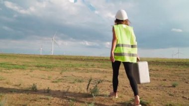Üniformalı ve kasklı kadın profesyonel ekoloji mühendisi elinde özel ekipmanlarla güzel bir gökyüzü ve saha arka planında yel değirmenine hizmet ediyor. Alternatif enerji kavramı. Yavaş çekim.