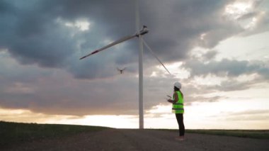 Üniformalı ve miğferli profesyonel ekoloji mühendisi güzel günbatımı arka planında rüzgar türbininde çalışan bir dron için uzaktan kumanda tutuyor. Elektrik enerjisine alternatif. Yavaş çekim.