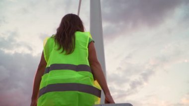 Kadın profesyonel ekoloji mühendisi elinde özel ekipmanlarla güzel bir günbatımı arka planında yel değirmenine hizmet ediyor. Alternatif enerji kavramı. Yavaş çekim.