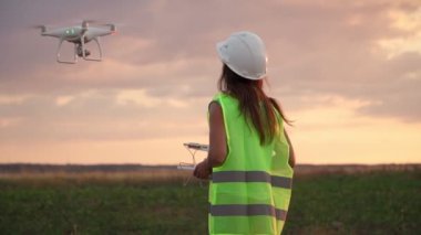 Üniformalı ve kasklı kadın ekoloji mühendisi elinde joystick ile yel değirmeninde çalışan uçan dronu kontrol ediyor. Elektrik enerjisine alternatif. Yavaş çekim.