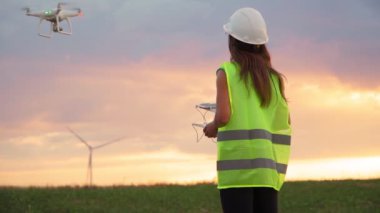 Üniformalı ve kasklı kadın ekoloji mühendisi elinde joystick ile yel değirmeninde çalışan uçan dronu kontrol ediyor. Elektrik enerjisine alternatif. Yavaş çekim.