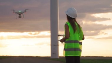 Üniformalı ve kasklı kadın ekoloji mühendisi elinde joystick ile yel değirmeninde çalışan uçan dronu kontrol ediyor. Elektrik enerjisine alternatif.