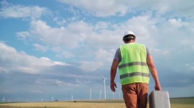 Üniformalı ve kasklı profesyonel ekoloji mühendisi elinde özel ekipmanlarla güzel bir arka planda yel değirmenine hizmet ediyor. Alternatif enerji ve rüzgar santrali. Yavaş çekim.