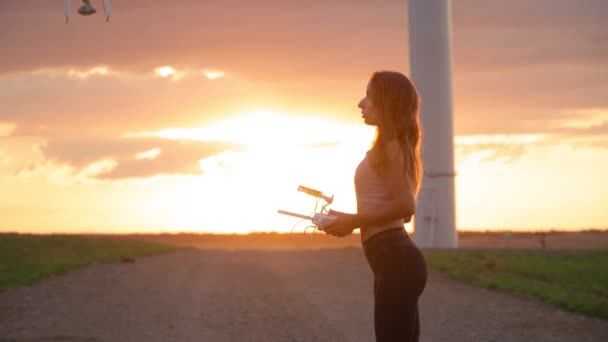 穿着运动服 手持遥控器的性感瘦小女人在美丽的日落和风车的背景下举起了无人机 慢动作 — 图库视频影像