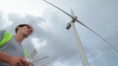 Üniformalı ve miğferli Uzman Ekoloji Mühendisi, yel değirmenlerinin arka planında çalışan insansız hava aracı için uzaktan kumanda tutuyor. Rüzgâr çiftliği servisi. Elektrik enerjisine alternatif.