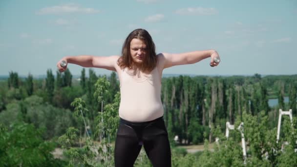 Hravý tlustý muž s dlouhými vlasy dělá gymnastiku pro hubnutí venku. Vtipný chlap s nadváhou zobrazující dívku zapojenou do fitness s činkami v parku. Zpomalený pohyb