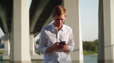 Beyaz tişörtlü yakışıklı genç adam şehir parkında oturuyor ve arkadaşlarıyla sosyal ağlarda iletişim kurarken cep telefonu kullanıyor ve gülüyor. Uzak iletişim