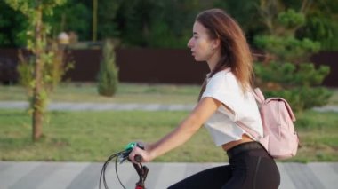 Güzel genç bir kadın güneşli bir yaz gününde şehir parkında bisiklet sürüyor. Sırtında pembe bir sırt çantası olan bisikletçi kız. Yavaş çekim.