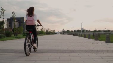 Güzel bir genç kadın bir yaz günü şehir parkında bisiklet sürüyor. Pembe sırt çantalı bisikletçi kadın. Arkadan bak. Yavaş çekim.