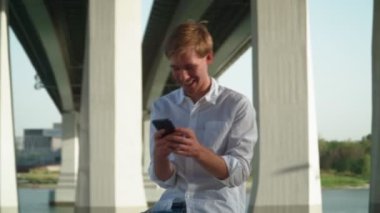 Beyaz tişörtlü yakışıklı genç adam şehir parkında oturuyor ve arkadaşlarıyla sosyal ağlarda iletişim kurarken cep telefonu kullanıyor ve gülüyor. Uzak iletişim