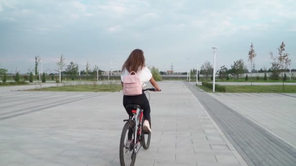 美しい若い女性は夏の日に都市公園で自転車に乗っています ピンクのバックパックを持つサイクリストの女の子 バックビュー スローモーション — ストック動画