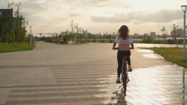 Güzel bir genç kadın bir yaz günü şehir parkında bisiklet sürüyor. Günbatımı arka planında pembe sırt çantalı bisikletçi kadın. Arkadan bak. Yavaş çekim.