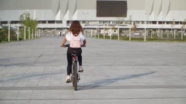 Güzel bir genç kadın bir yaz günü şehir parkında bisiklet sürüyor. Pembe sırt çantalı bisikletçi kız. Arkadan bak. Yavaş çekim.