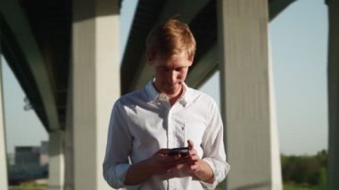 Beyaz tişörtlü yakışıklı genç adam şehir parkında oturuyor ve arkadaşlarıyla sosyal ağlarda iletişim kurarken cep telefonu kullanıyor ve gülüyor. Uzak iletişim