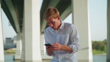 Beyaz tişörtlü yakışıklı genç adam şehir parkında oturuyor ve arkadaşlarıyla sosyal ağlarda iletişim kurarken cep telefonu kullanıyor ve gülüyor. Uzak iletişim