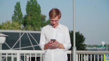 Beyaz gömlekli yakışıklı genç adam cep telefonu kullanarak gülümsüyor. Sosyal medya aracılığıyla telefonla konuşmaktan hoşlanıyor. Yavaş çekim