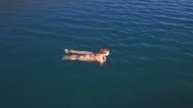 Hava görüntüsü. Mayo giymiş ince vücutlu güzel bir kadın kristal denizde yüzüyor.
