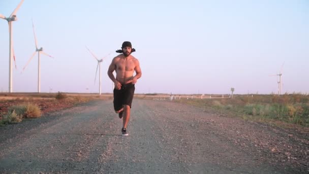 Musculoso atlético hombre árabe con barba corriendo en el campo contra molino de viento telón de fondo — Vídeos de Stock
