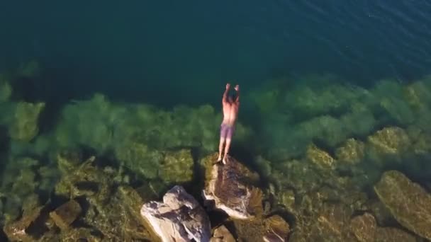 Luchtdrone gespierde man springt van rots naar helder blauw water in meer en zwemt — Stockvideo