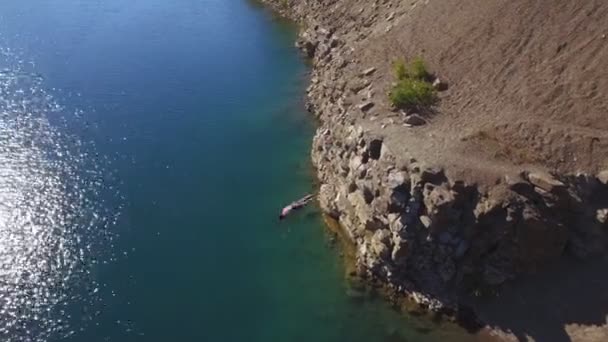 Aerial view. muscular man jumps off cliff into crystal clear sea water and swim — Stock Video