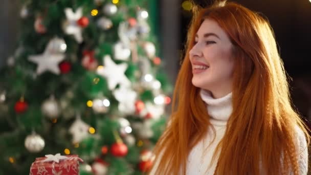 Hermosa mujer pelirroja feliz sentado fondo árbol de Navidad y sonriendo — Vídeo de stock