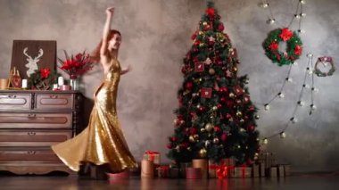 Altın gece elbisesi içinde güzel bir kadın Noel ağacının etrafında dönüyor ve dans ediyor.