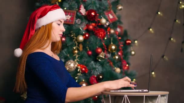 Woman in Santa hat works laptop and types on keyboard background Christmas tree. — Stock Video