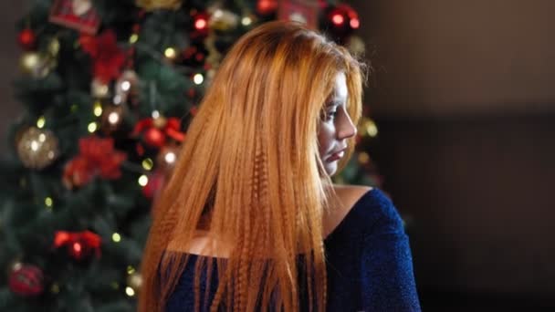 Mooie roodharige vrouw kijkt naar camera tegen de achtergrond kerstboom — Stockvideo